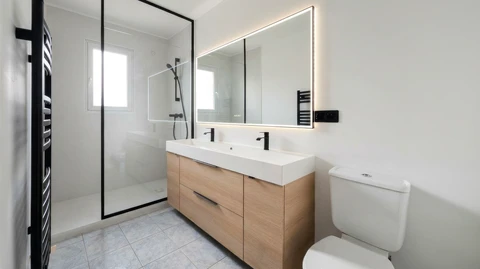 Salle de bain moderne épurée avec douche à l'italienne, paroi en verre à cadre noir, meuble vasque double suspendu en bois clair avec plan vasque blanc intégré, robinetterie noire mate, grand miroir LED rétroéclairé, WC à réservoir apparent, sèche-serviettes électrique noir, carrelage grès cérame gris clair, murs blancs